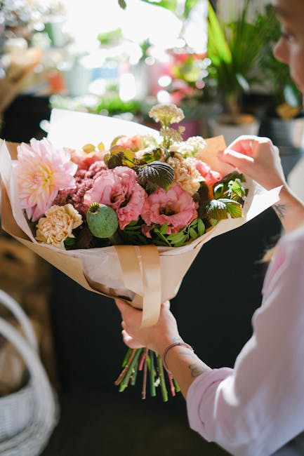 A person holding a large bouquet of fresh flowers, including pink peonies, white roses, and purple hydrangeas, wrapped in beige craft paper with a satin ribbon. The bouquet features a mix of blooms an