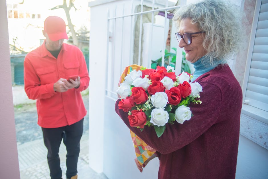 A woman with curly gray hair, wearing glasses, a light blue turtleneck, and a maroon cardigan, holds a vibrant bouquet of fresh roses in red and white, wrapped in patterned paper, during a flower deli
