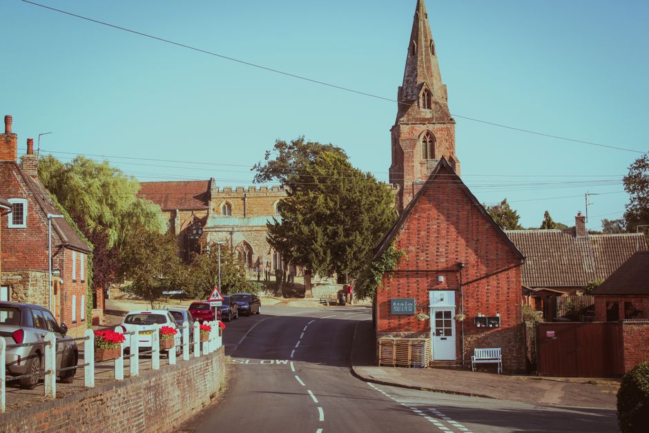 A picturesque scene of a small village street in Great Bookham, KT23, featuring a red brick building with a white door and a small white bench outside. In the foreground, there are white-painted raili