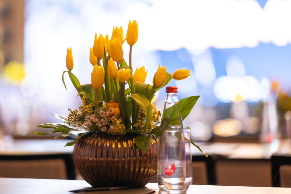 A floral arrangement featuring tall, vibrant yellow tulips with closed buds and green stems, arranged in a dark, ribbed glass vase on a light-colored table. The bouquet includes small white and yellow