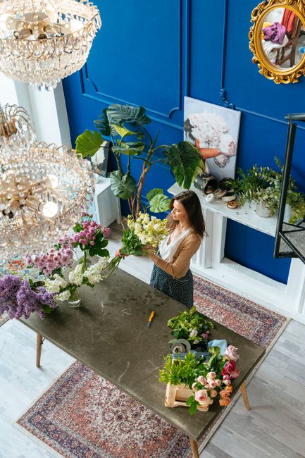 A woman in a beige top and dark skirt arranges a vibrant bouquet of white and yellow flowers, including roses and lilies, on a dark, rustic table inside a stylish floral shop. The workspace is filled 