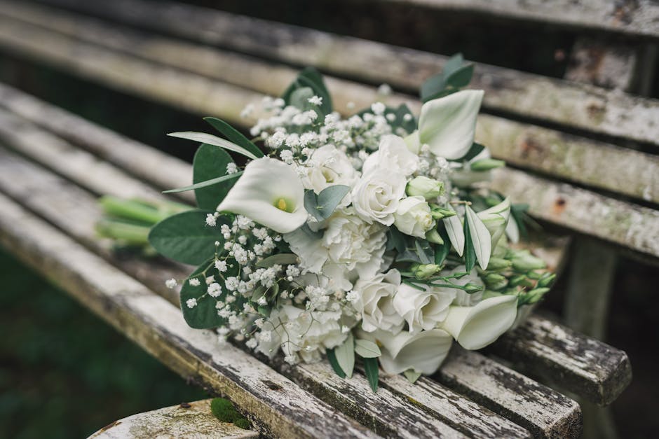 A bouquet of fresh white flowers resting on a weathered wooden bench outdoors, featuring elegant calla lilies, roses, and freesias accented with eucalyptus leaves and baby's breath. The flowers displa