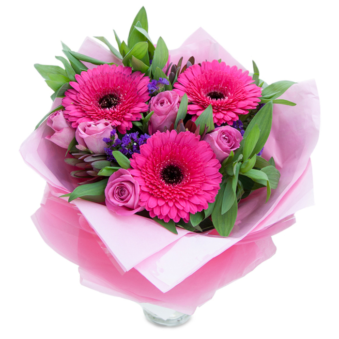 A tender, springlike bouquet pictured against a white field, composed with the kind of attention to balance and texture you expect from an experienced florist. Three fuchsia Gerbera daisies command the eye, their layered petals opening wide around pronounced dark centres that offer a pleasing, almost graphic contrast. Circling them are several pale lavender-pink roses-buds that unfurl slowly with velvety petals-softening the overall effect and lending a romantic, intimate note. Small stems of purple statice or limonium are tucked through the arrangement, their tiny clustered flowers adding a papery texture and a cool purple accent that plays off the warmer pink tones. The greenery is generous and glossy; broad salal or eucalyptus leaves create a rich, green backdrop and give the bouquet a fresh, just-cut-from-the-garden feel. For presentation the blooms are nestled in soft matte pale-pink tissue and wrapped with a layer of translucent, slightly crinkled fuchsia-pink cellophane that shimmers discreetly in reflected light; the careful folds of the wrap demonstrate the florist's craft. At the lower edge the rim of a clear glass vase is visible, indicating the arrangement is ready for immediate display on a sideboard or dining table. Under gentle daylight the roses emit a subtle sweet perfume while the gerberas contribute a faint, green-fresh scent, together creating an inviting sensory impression. This composition is perfect for marking birthdays, anniversaries, Mother's Day or a thoughtful surprise for a neighbour in Great Bookham village-an approachable, elegant bouquet designed to brighten a room and lift spirits.